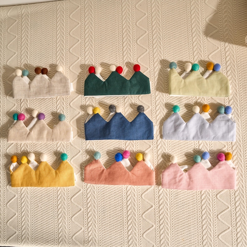 May include: Nine fabric crowns in various colors, each with three peaks and topped with colorful pom-poms. The crowns are arranged on a cream-colored, textured surface. Colors include green, blue, yellow, pink, and neutral tones.