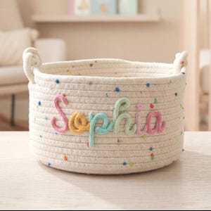 May include: A round, off-white rope basket with colorful speckles and two rope handles. The name "Sophia" is embroidered in pastel colors across the front. This basket is ideal for nursery storage.