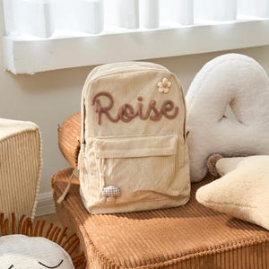 May include: A beige corduroy backpack with a brown embroidered name "Roise" and a small flower and mushroom applique.