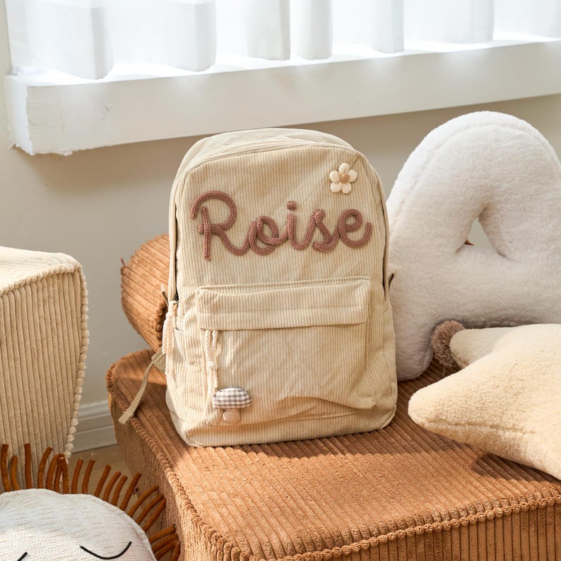 May include: A beige corduroy backpack with a brown embroidered name "Roise" and a small flower and mushroom applique.