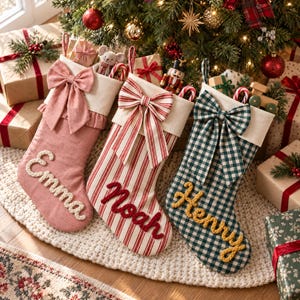 Op de afbeelding: Drie kerstsokken met crèmekleurige boorden en grote strikken. De eerste is effen roze met de naam Emma, de tweede heeft rode en witte strepen met de naam Noah, en de derde heeft een groen-wit ruitpatroon met de naam Henry.