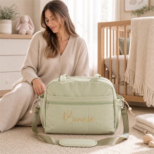 May include: A sage green changing bag featuring a horizontal quilted pattern and a zippered front pocket. The name Bonnie is embroidered in gold cursive script on the front. The bag includes a matching adjustable shoulder strap with a padded section.