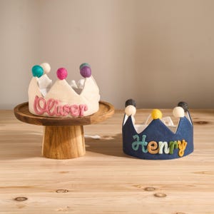 May include: Two handmade fabric crowns. One is cream-colored with the name "Oliver" in pink script, and the other is navy blue with the name "Henry" in colorful letters. Both crowns have decorative pom-poms.