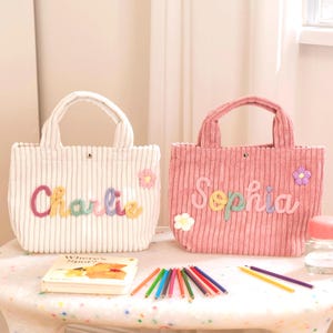 May include: Two corduroy tote bags, one white and one pink, with embroidered names and floral accents. The white bag says "Charlie" in colourful letters, and the pink bag says "Sophia." A children's book and coloured pencils are on the table.