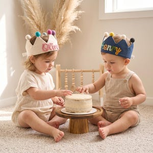 May include: Two babies wearing fabric crowns with colorful pom-poms and names on them, sitting on a carpet. They are reaching for a small cake on a wooden stand. One crown says "Olive" and the other says "Henry".