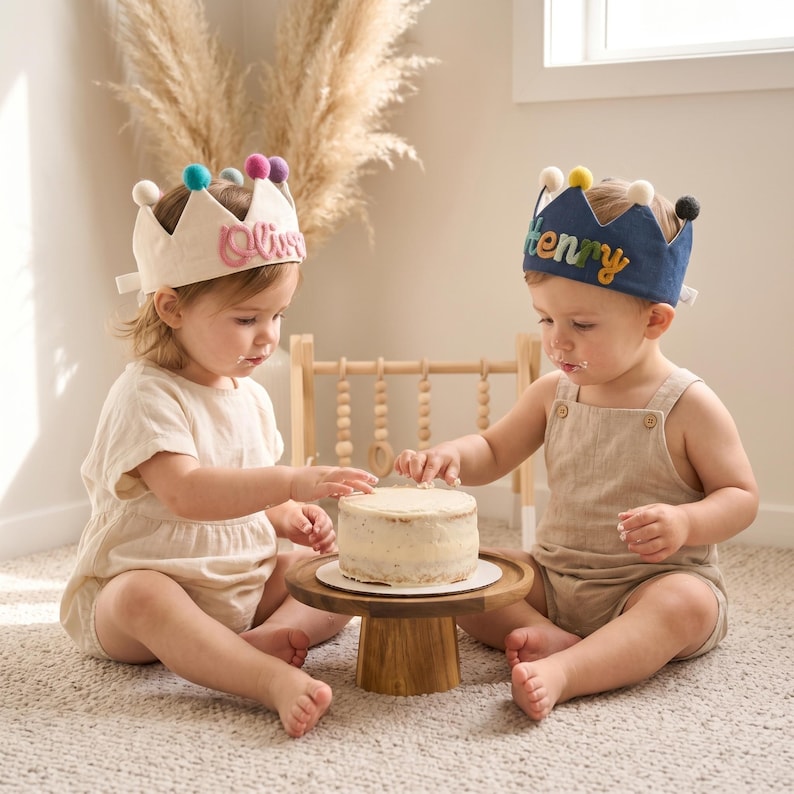 May include: Two babies wearing fabric crowns with colorful pom-poms and names on them, sitting on a carpet. They are reaching for a small cake on a wooden stand. One crown says "Olive" and the other says "Henry".