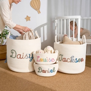 Op de afbeelding: Vier crèmekleurige touw opbergmanden met geborduurde namen: "Daisy", "Louise", "Jackson" en "Daniel". De manden bevatten speelgoed en kleding. Een persoon stopt kleding in de grootste mand. Een baby zit op de achtergrond in een wieg.