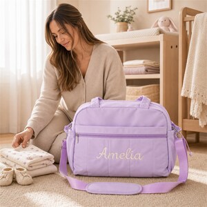 Peut inclure: Un sac à langer violet clair avec un motif matelassé horizontal, une poche frontale zippée et le prénom Amelia brodé dans une police cursive claire. Le sac comprend deux poignées supérieures et une longue bandoulière réglable.