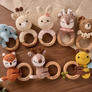 May include: A collection of handmade crochet baby rattles in various animal shapes, including an elephant, giraffe, bunny, deer, bear, fox, and bee. Each rattle features a wooden ring and is personalized with a name and date. The rattles are in shades of blue, beige, brown, orange, and yellow.