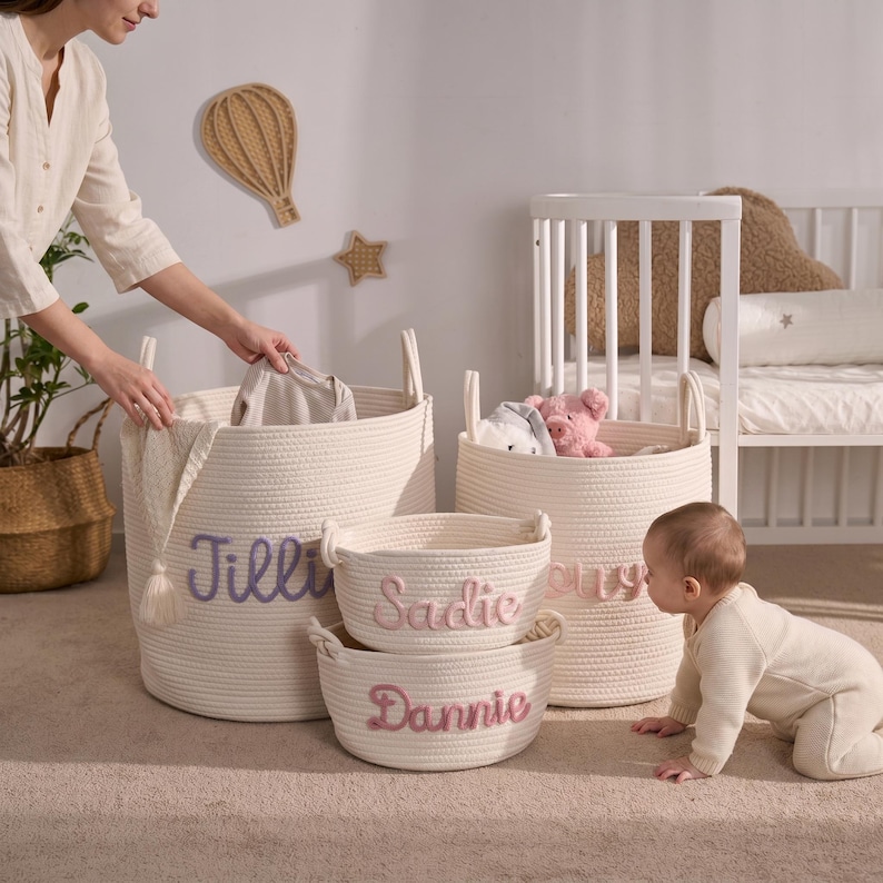 May include: A set of four off-white woven rope storage baskets with integrated handles. The baskets feature personalized names embroidered in soft pink and lavender yarn on the front. The collection includes two large upright baskets and two smaller, shallower round baskets.