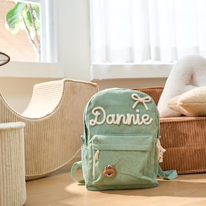 May include: A green corduroy backpack with a white embroidered name "Dannie" and a white bow. The backpack has a brown bear charm and a small pocket on the front.