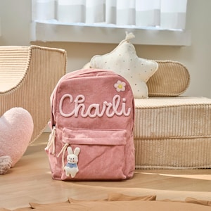May include: A pink corduroy backpack with a white bunny charm and the name "Charli" embroidered on the front. The backpack has a small flower patch on the top.