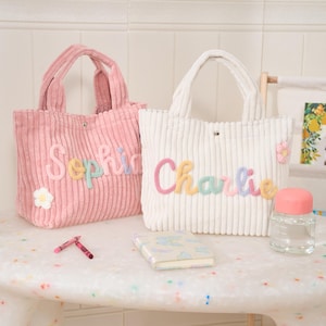 Op de afbeelding: Twee corduroy draagtassen, één roze en één witte, met geborduurde namen en bloemaccenten. De roze tas heeft de tekst "Sophia" en de witte tas "Charlie". Beide tassen hebben bijpassende handvatten.