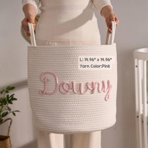 May include: A white woven rope storage basket with two handles, featuring the name Downy embroidered in pink yarn on the front. The basket measures 14.96 inches by 14.96 inches, with the yarn color specified as pink on the attached label.