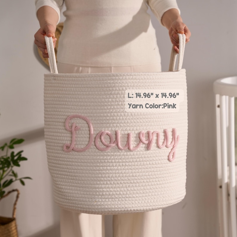 May include: A white woven rope storage basket with two handles, featuring the name Downy embroidered in pink yarn on the front. The basket measures 14.96 inches by 14.96 inches, with the yarn color specified as pink on the attached label.