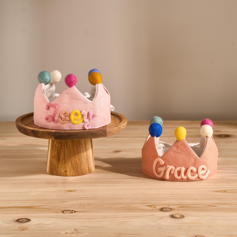 May include: Two pink fabric crowns with colorful pom-poms. One crown is on a wooden cake stand and has the name "Zoey" embroidered in colorful thread. The other crown has the name "Grace" embroidered in white thread.