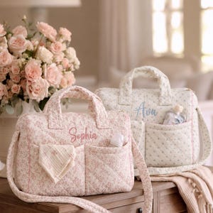Peut inclure: Deux sacs à langer matelassés avec poignées et bandoulières. Le sac avant est rose avec un motif floral et le prénom Sophia brodé en rose. Le sac arrière est blanc avec des motifs floraux bleus et le prénom Ava brodé en bleu.