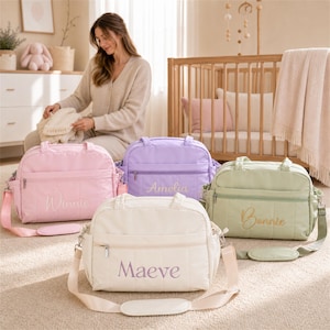 May include: Four rectangular quilted changing bags with adjustable shoulder straps and front zippered pockets. The bags are coloured light pink, lavender, sage green, and cream, each featuring embroidered names: Winnie, Amelia, Bonnie, and Maeve.