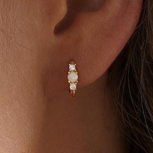 May include: Gold hoop earring featuring an opal stone and two smaller clear stones. The earring is displayed on an ear, highlighting the design and colour. A small, delicate piece of jewellery.