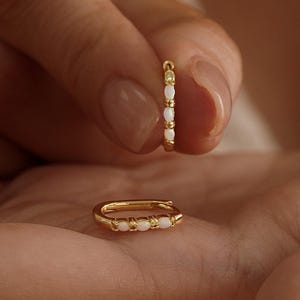 May include: A pair of gold hoop earrings featuring small, white, oval-shaped stones set along the front edge. One earring is held upright between fingers, while the other rests horizontally in the palm. The text ArtisanalEleCreate is visible.