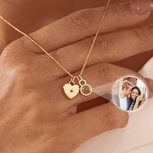 May include: A gold necklace featuring a heart-shaped pendant and a circular pendant. The heart pendant has a small star design. The round pendant seems to contain a small photo. The necklace is worn around the neck.