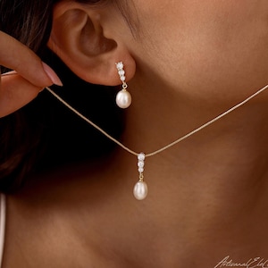 May include: A gold necklace and earring set featuring a delicate box chain and drop earrings. Each piece displays three small, clear, round-cut stones arranged vertically above a single, smooth, white, oval-shaped pearl pendant.