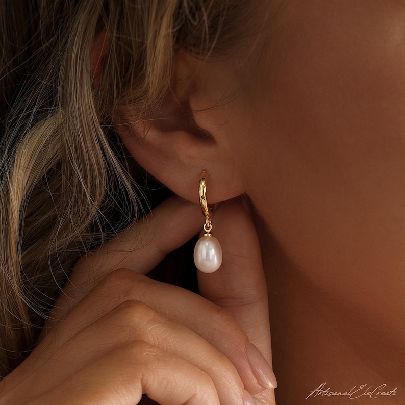 May include: A gold hoop earring featuring a single, smooth, teardrop-shaped white pearl pendant. The earring is crafted from polished gold metal, with the pearl suspended from the base of the hoop, creating a simple and elegant drop design.