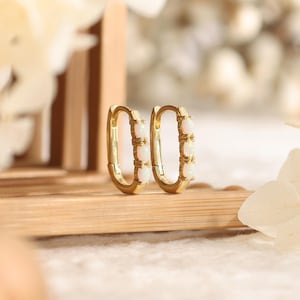 May include: A pair of gold hoop earrings featuring an elongated oval shape. Each earring is accented with three small, round, iridescent white opal stones set vertically along the front-facing outer edge of the polished gold metal band.