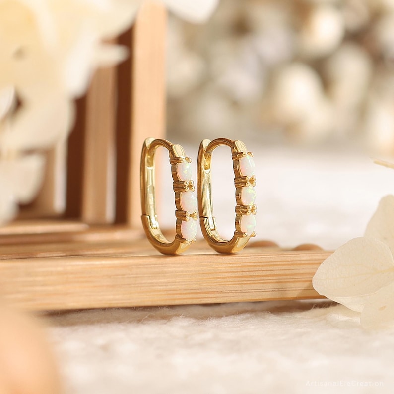 May include: A pair of gold hoop earrings featuring an elongated oval shape. Each earring is accented with three small, round, iridescent white opal stones set vertically along the front-facing outer edge of the polished gold metal band.