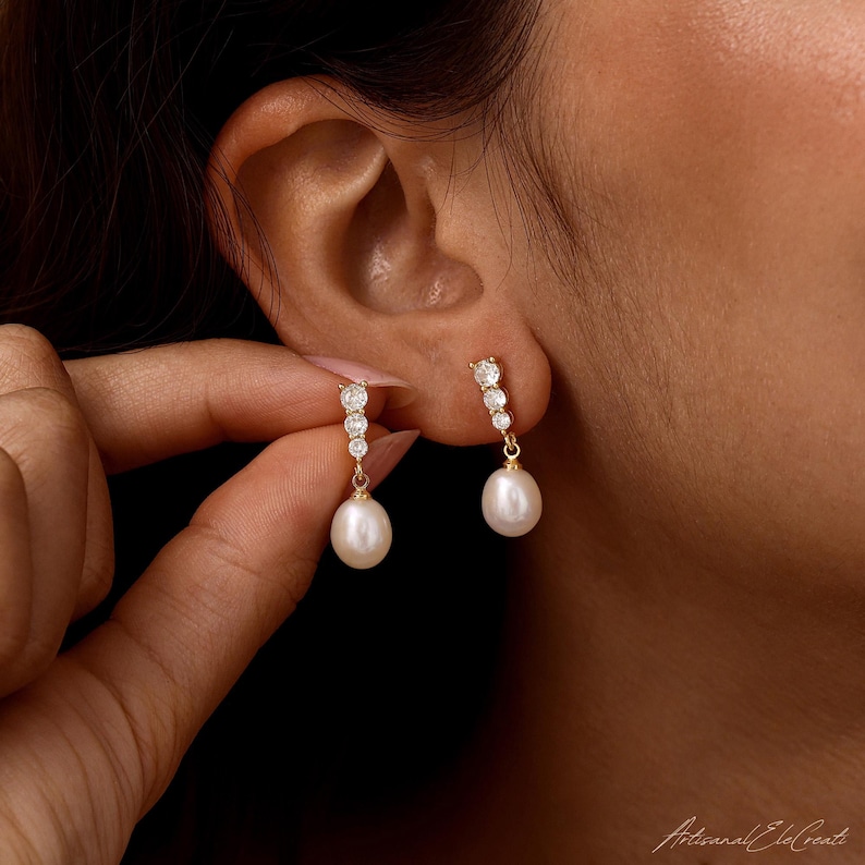 May include: A pair of gold drop earrings featuring three small, clear, round-cut stones arranged vertically, terminating in a single, smooth, white, teardrop-shaped pearl. The earrings are suspended from a thin gold post and secure with a back.