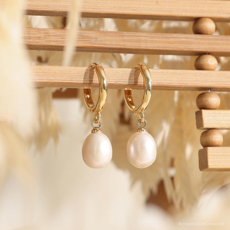 May include: A pair of gold hoop earrings featuring dangling, teardrop-shaped white pearls. The earrings are suspended from a light-colored wooden structure. The metal hoops are polished and reflective, while the pearls have a smooth, lustrous finish.