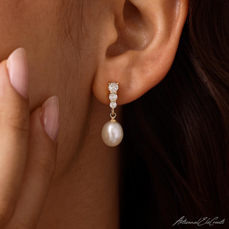 May include: A gold drop earring featuring a vertical row of three small, round, clear stones set in gold, terminating in a single, smooth, white, teardrop-shaped pearl that hangs from the base of the stone setting, secured to the earlobe.