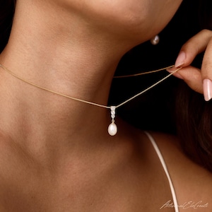 May include: A gold chain necklace featuring a pendant with three small clear stones arranged vertically above a single teardrop-shaped white pearl. The necklace is worn around the neck, and a matching pearl earring is visible near the jawline.