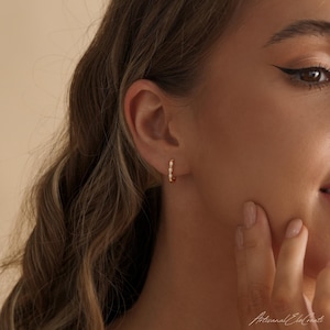 May include: A small gold hoop earring featuring a series of tiny, round, white opalescent stones set along the outer edge. The earring is worn in the earlobe, showcasing a polished metallic finish and a delicate, circular design with multiple stones.