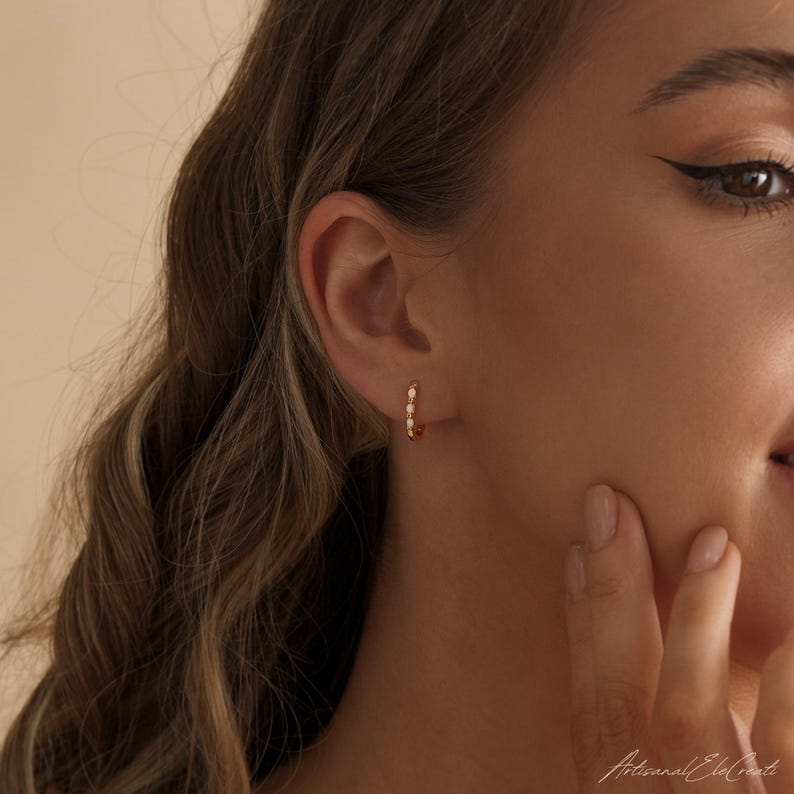 May include: A small gold hoop earring featuring a series of tiny, round, white opalescent stones set along the outer edge. The earring is worn in the earlobe, showcasing a polished metallic finish and a delicate, circular design with multiple stones.