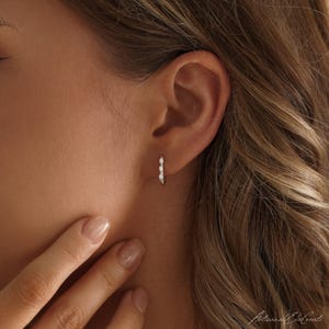 May include: A small hoop earring featuring three round white opal stones set in a thin metallic band. The earring is worn in the earlobe, showcasing a delicate design with a polished finish and a secure clasp mechanism for everyday wear.