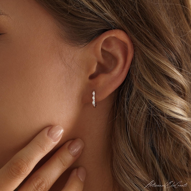 May include: A small hoop earring featuring three round white opal stones set in a thin metallic band. The earring is worn in the earlobe, showcasing a delicate design with a polished finish and a secure clasp mechanism for everyday wear.