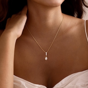 May include: A gold chain necklace featuring a vertical drop pendant. The pendant consists of two small, clear, round-cut stones set in gold, terminating in a single, white, oval-shaped pearl drop. The necklace rests against the collarbone area.