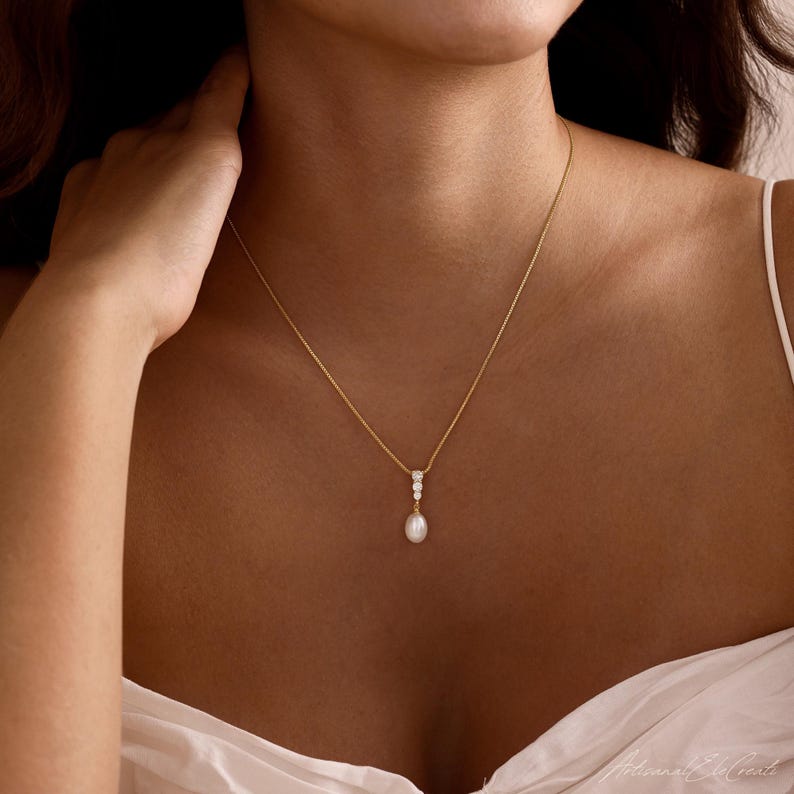May include: A gold chain necklace featuring a vertical drop pendant. The pendant consists of two small, clear, round-cut stones set in gold, terminating in a single, white, oval-shaped pearl drop. The necklace rests against the collarbone area.
