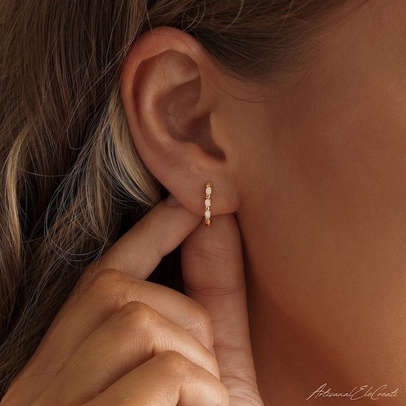 May include: A small gold hoop earring featuring three evenly spaced oval white opals set along the front of the band. The earring is secured in the earlobe, showcasing a polished metallic finish and a delicate, refined design with clear stones.