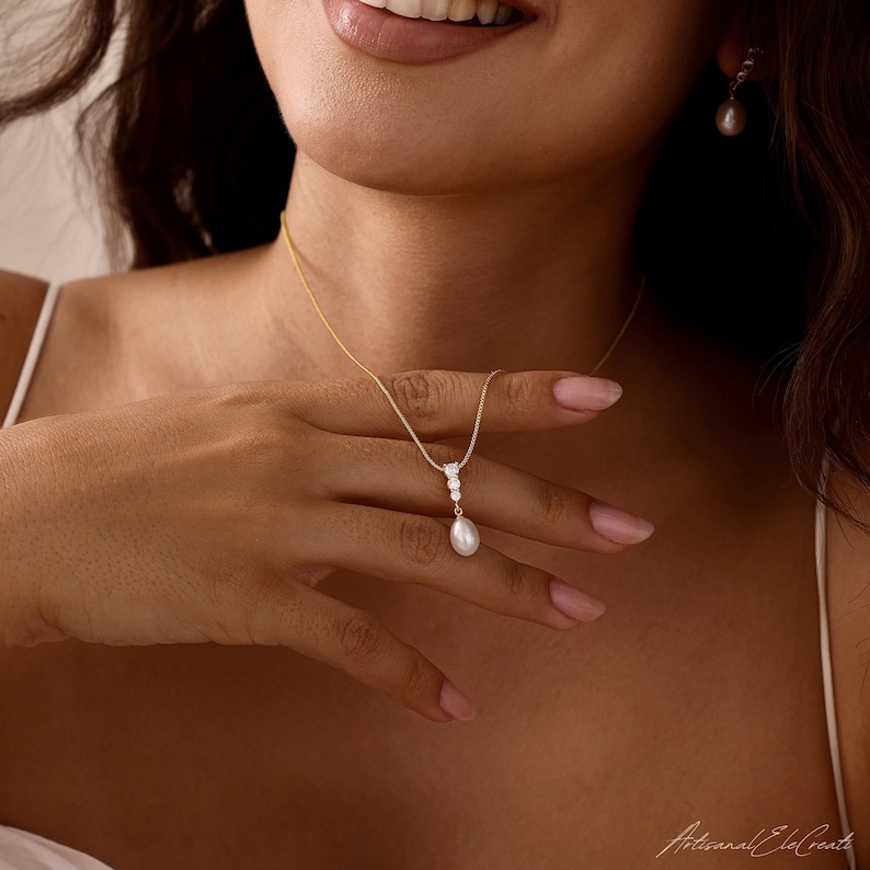 May include: A gold chain necklace featuring a pendant with three small clear stones and a single white teardrop pearl. The jewelry set includes a matching gold earring with a single white teardrop pearl, creating a coordinated look with a delicate design.