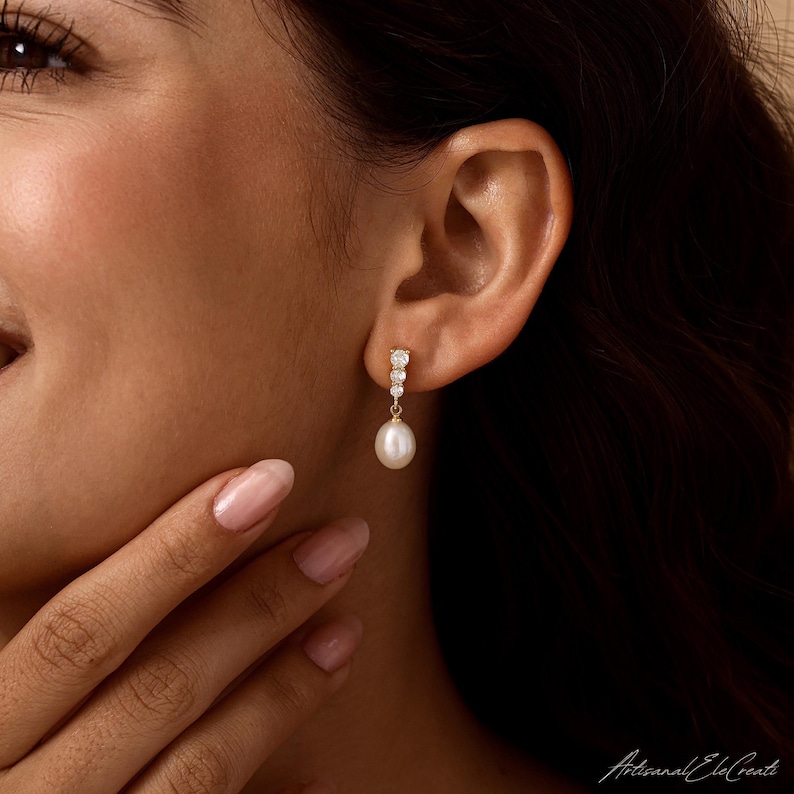 May include: A gold drop earring featuring a vertical row of three small, clear, round-cut stones leading to a single, smooth, white, teardrop-shaped pearl that hangs delicately from the base of the metal setting against the earlobe.