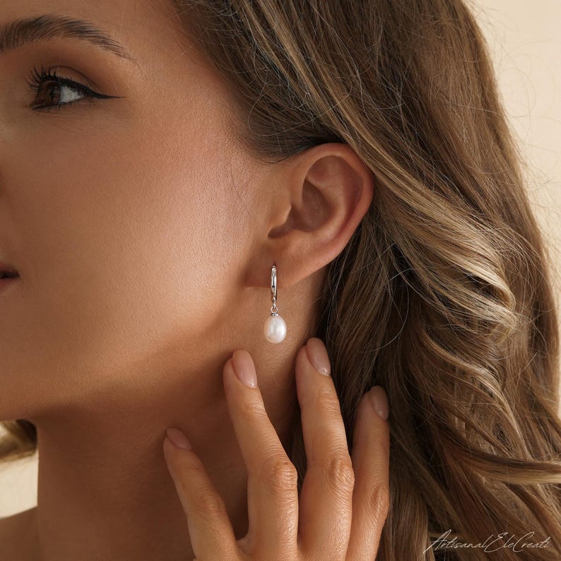 May include: A silver hoop earring featuring a single, smooth, white teardrop-shaped pearl dangling from the bottom. The earring is secured through the earlobe, showcasing a polished metallic finish and a classic, simple design with a lustrous pearl.