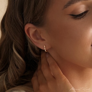 May include: A small gold hoop earring featuring a series of tiny, round, white opal gemstones set along the front curve. The earring is worn in the earlobe, showcasing a polished metallic finish and a delicate, circular design structure.