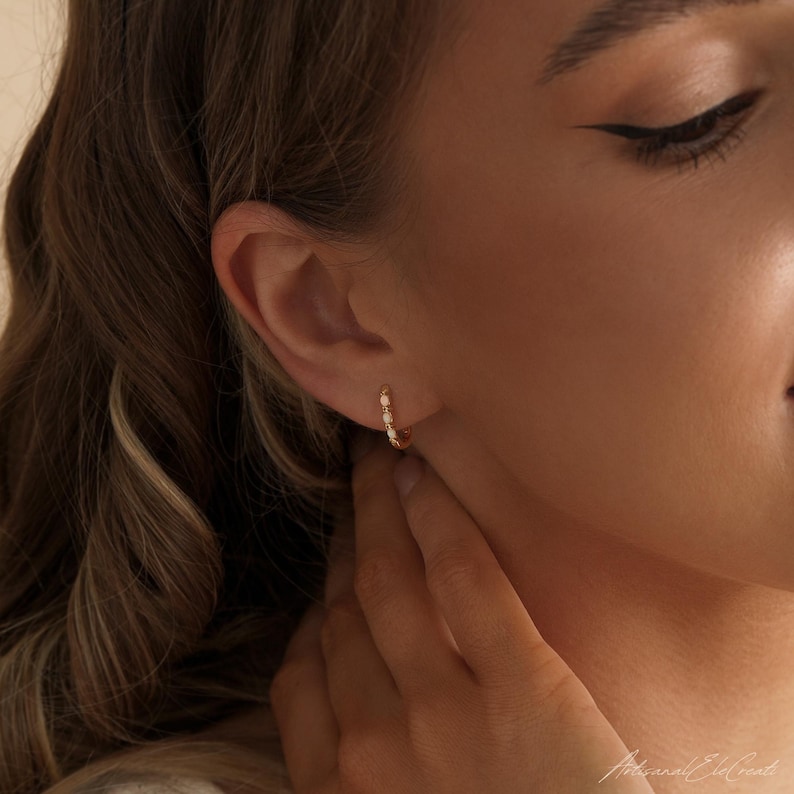 May include: A small gold hoop earring featuring a series of tiny, round, white opal gemstones set along the front curve. The earring is worn in the earlobe, showcasing a polished metallic finish and a delicate, circular design structure.