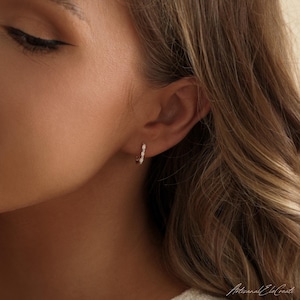 May include: A small silver hoop earring featuring three oval-shaped white opal stones set in a vertical row along the front of the hoop. The earring is worn in the earlobe, showcasing a polished metallic finish and iridescent stone accents.