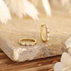 May include: A pair of gold hoop earrings featuring three small, oval-shaped white opals set along the front edge of each hoop. One earring is lying flat while the other stands upright, showcasing the polished gold metal and the iridescent stones.
