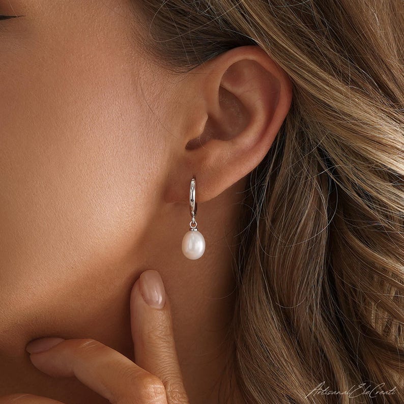 May include: A silver hoop earring featuring a single, smooth, teardrop-shaped white pearl dangling from the bottom. The earring is secured through the earlobe, showcasing a polished metallic finish on the hoop and a lustrous surface on the pearl.