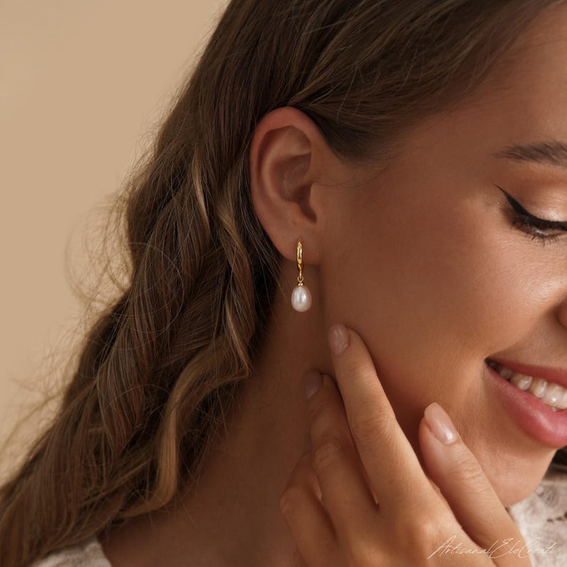 May include: A gold hoop earring featuring a single, small, white, teardrop-shaped pearl pendant. The earring is worn in the earlobe, showcasing a polished gold metal finish on the hoop and a smooth, lustrous surface on the pearl drop component.