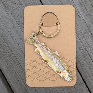 Könnte beinhalten: Ein fischförmiger Schlüsselanhänger mit einem bronzefarbenen Ring und Verschluss. Der Fisch ist mit realistischen Details bemalt, einschließlich schillernder Schuppen und Flossen. Der Schlüsselanhänger ist an einer braunen Karte mit Wellenmuster befestigt.